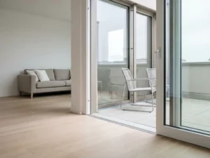 Offene Glas-Schiebetür mit Blick auf hellen Balkon, Greige-Sofa, Holzboden und zwei schlanke Terrassenstühle in minimalistischer Wohnung.