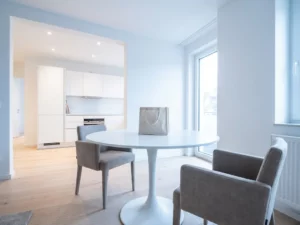 Minimalistisches Hannoveraner Apartment mit rundem weißem Tulpen-Esstisch, zwei greigefarbenen Stühlen und einer Stofftasche auf der Tischplatte.