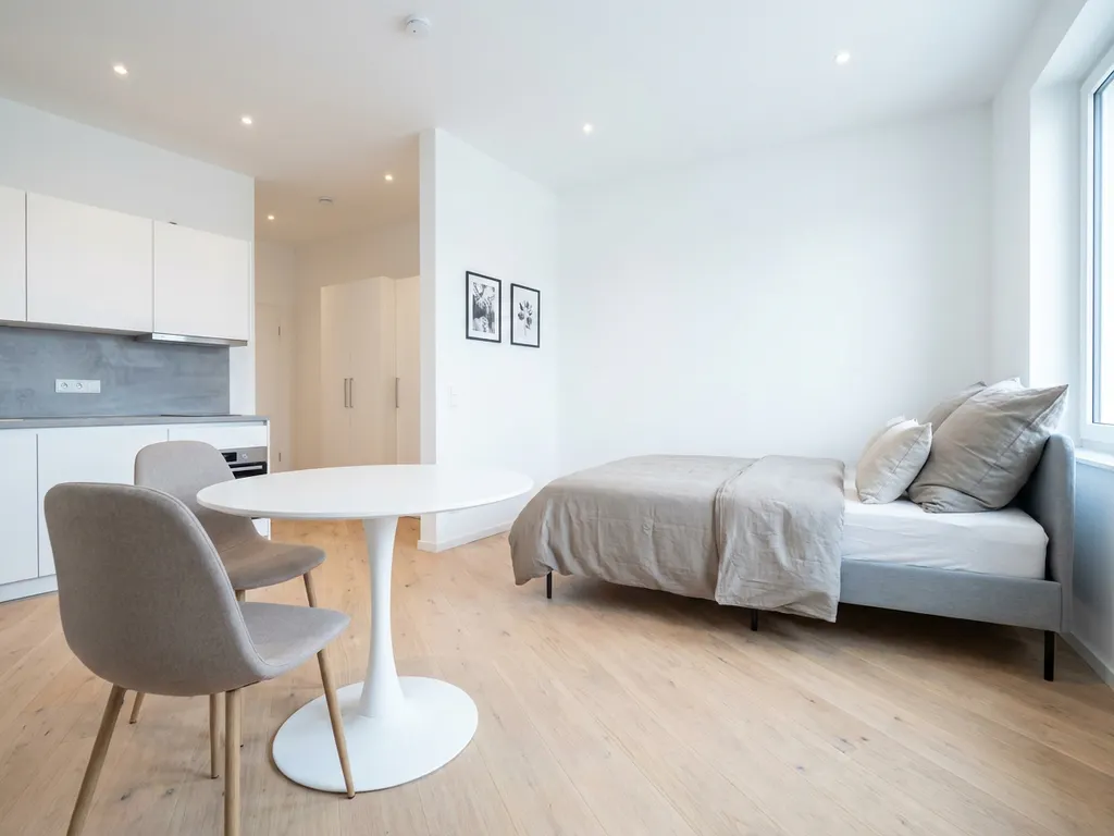 Minimalistisches Ein-Zimmer-Apartment in Hannover mit greigem Bett, Tulpen-Esstisch und hellem Holzboden.