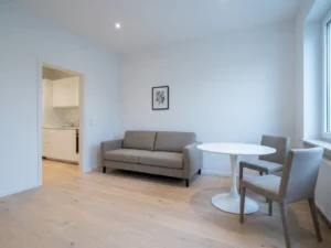 Minimalistisches Wohnzimmer in Hannover mit hellem Parkettboden, graubeiger Couch und weißem Tulpen-Esstisch vor offener Küche.