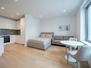 Helles Mikro-Apartment mit Parkettboden, greigem Sofa, weißem Tulip-Esstisch und offener Küche in Weiß und Grau.