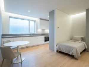 Minimalistisches Apartment mit hellem Eichenparkett, weißer Einbauküche, grauer Steinarbeitsplatte und rundem Tulpen-Esstisch mit greigen Polsterstühlen.
