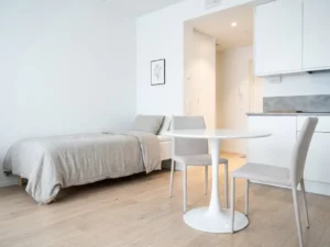 Minimalistisch eingerichtetes Apartment mit Einzelbett, rundem Tulip-Tisch und hellem Parkettboden in Greige- und Weißtönen.