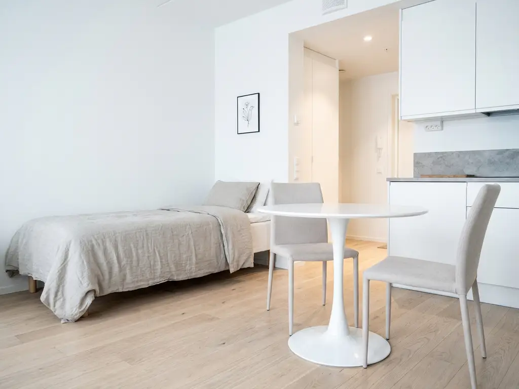 Minimalistisch eingerichtetes Apartment mit Einzelbett, rundem Tulip-Tisch und hellem Parkettboden in Greige- und Weißtönen.