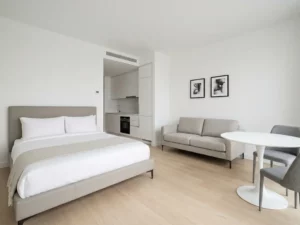 Minimalistisch eingerichtetes Apartment mit greigem Bett, weißer Küche, Tulpen-Esstisch und hellem Eichenholzboden.