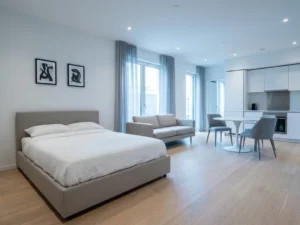 Minimalistisch eingerichtetes Apartment mit greigebeigem Bett, weißem Tulpen-Esstisch und hellen Holzdielen.