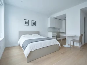 Minimalistisch eingerichtetes Apartment mit greigem Bettgestell, rundem Tulpen-Esstisch und hellen Holzböden in ruhiger Wohn-Schlaf-Kombination.