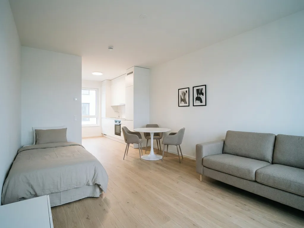 Minimalistisch eingerichtetes Apartment mit greigem Bett, Sofa, rundem Tulpen-Esstisch und weißer Einbauküche auf hellem Parkettboden.