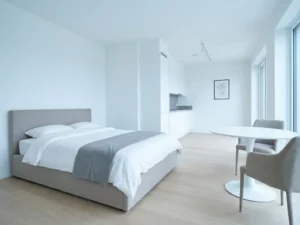 Minimalistisch eingerichtetes Apartment mit greigem Bettgestell, rundem Tulpen-Esstisch und hellem Eichenholzboden.