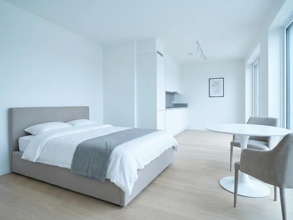 Minimalistisch eingerichtetes Apartment mit greigem Bettgestell, rundem Tulpen-Esstisch und hellem Eichenholzboden.