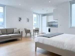 Minimalistisch eingerichtete Wohnung mit greigem Bettgestell, Zweisitzer-Sofa, rundem Tulpen-Esstisch und weißen Küchenschränken auf hellem Eichenparkett.