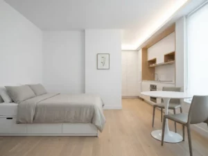 Minimalistisch eingerichtetes Apartment mit Einzelbett, rundem Tulpen-Esstisch, zwei Greige-Stühlen und weißer Einbauküche auf hellem Parkettboden.