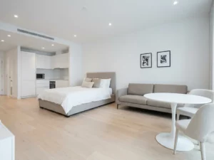 Minimalistisch eingerichtetes Apartment mit greigem Bettgestell, Zweisitzer-Sofa, rundem Tulpen-Esstisch und weißer Einbauküche auf hellem Parkettboden.