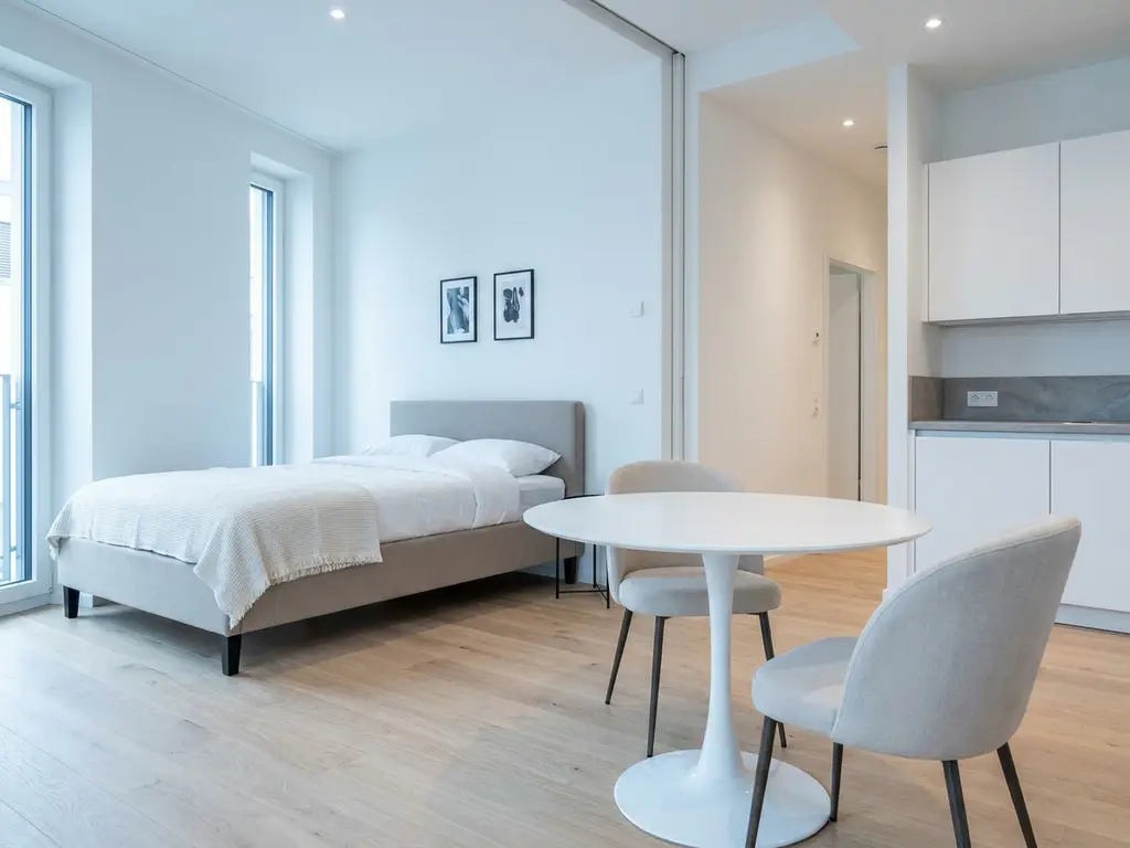 Minimalistisch eingerichtetes Apartment mit greigem Bettgestell, rundem Tulpen-Esstisch und hellem Eichenparkett.