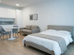 Minimalistisch eingerichtetes Apartment mit greigem Bett, Zweisitzer-Sofa, rundem Tulpen-Esstisch und hellen Holzdielen.
