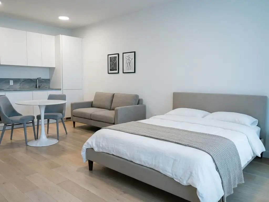 Minimalistisch eingerichtetes Apartment mit greigem Bett, Zweisitzer-Sofa, rundem Tulpen-Esstisch und hellen Holzdielen.