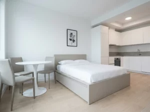 Minimalistisch eingerichtetes Apartment mit greigem Bettgestell, rundem Tulpen-Esstisch und hellen Eichenholzdielen.