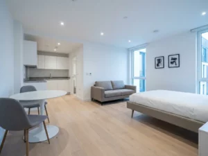 Minimalistisch eingerichtete offene Wohnung mit greigebeigem Sofa, Tulip-Esstisch, weißen Küchenfronten und hellem Eichenparkett.