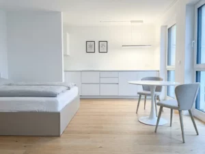 Minimalistisch eingerichtetes Apartment in Hannover mit greigem Bettgestell, rundem Tulpen-Esstisch und weißen Küchenfronten auf hellem Eichenparkett.