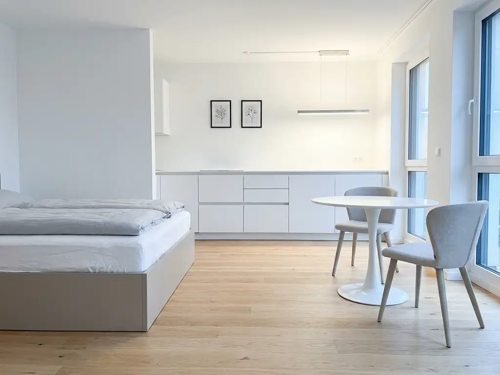 Minimalistisch eingerichtetes Apartment in Hannover mit greigem Bettgestell, rundem Tulpen-Esstisch und weißen Küchenfronten auf hellem Eichenparkett.