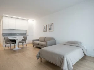Minimalistisch eingerichtetes Apartment mit Greige-Bett, weißer Tulpen-Esstisch, hellen Holzböden und weißen Küchenfronten.