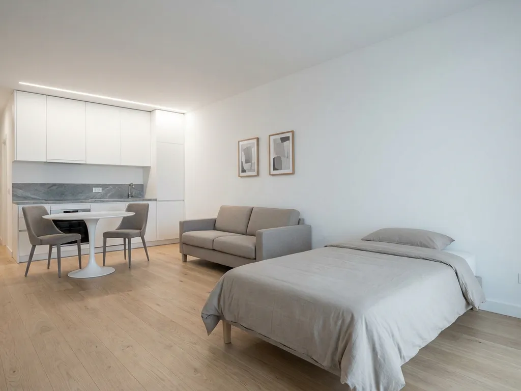 Minimalistisch eingerichtetes Apartment mit Greige-Bett, weißer Tulpen-Esstisch, hellen Holzböden und weißen Küchenfronten.