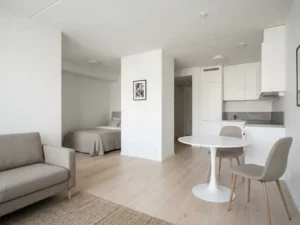Minimalistisch eingerichtete Wohnung mit hellem Parkettboden, weißer Küche, rundem Tulpen-Esstisch und graubeigem Sofa in neutraler Farbpalette.
