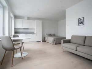 Minimalistisch eingerichtetes Apartment mit hellem Parkettboden, weißer Küche, rundem Tulip-Esstisch und greigefarbenem Sofa.