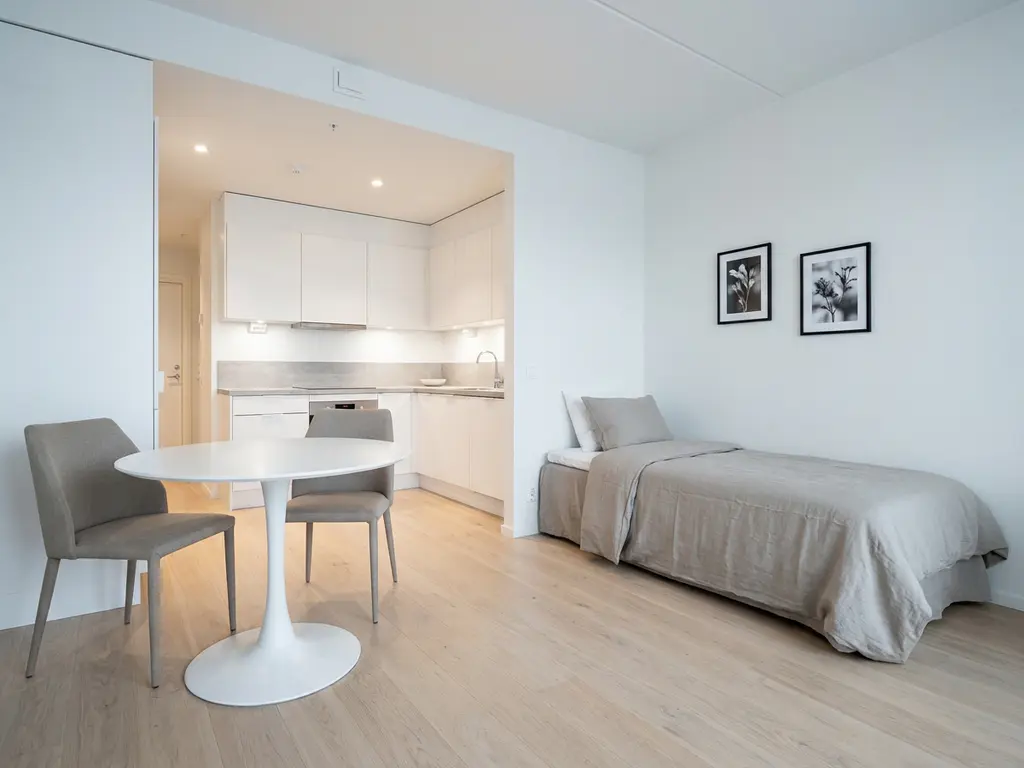 Minimalistisch möbliertes Apartment mit hellem Parkettboden, weißer Küche, rundem Tulpen-Esstisch und Einzelbett mit greigen Textilien.