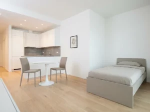 Minimalistisch eingerichtete Wohnung mit hellem Parkettboden, weißem Tulpen-Esstisch, greigefarbenen Stühlen und Bett sowie weißer Einbauküche.