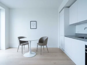 Minimalistisches Apartment mit hellem Parkettboden, weißer Einbauküche, grauer Steinarbeitsplatte und rundem Tulpen-Esstisch mit zwei greigen Polsterstühlen.