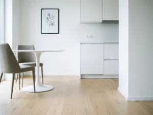 Leere minimalistische Wohnung mit hellem Parkettboden, weißem Tulpen-Esstisch und greigen Polsterstühlen vor weißer Einbauküche.
