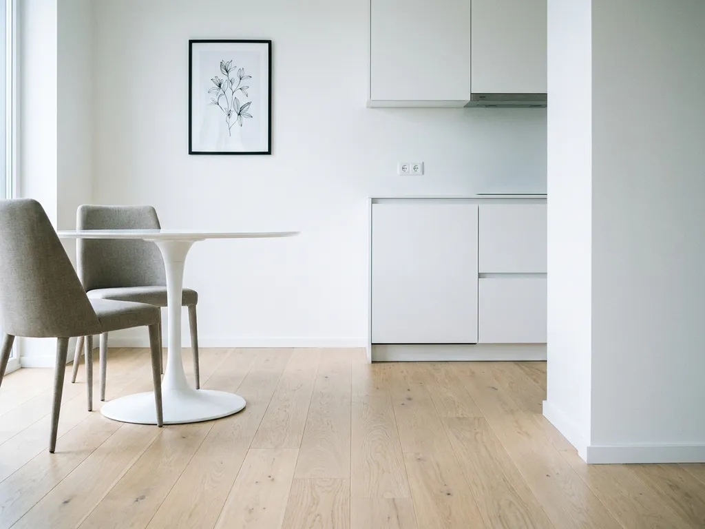 Leere minimalistische Wohnung mit hellem Parkettboden, weißem Tulpen-Esstisch und greigen Polsterstühlen vor weißer Einbauküche.