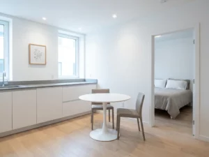 Minimalistisch eingerichtete Wohnung mit hellem Holzboden, weißer Küche, rundem Tulpen-Esstisch und zwei greige Stühlen.