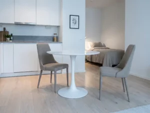 Minimalistisch eingerichtetes Apartment mit hellem Holzboden, weißem Tulpen-Esstisch, greige Stühlen und weißen Küchenschränken im Hintergrund.