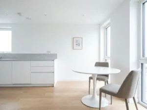 Minimalistisch eingerichtete Wohnung mit hellem Parkettboden, weißen Küchenschränken, grauer Arbeitsplatte und rundem Tulpen-Esstisch mit zwei greigen Stühlen.