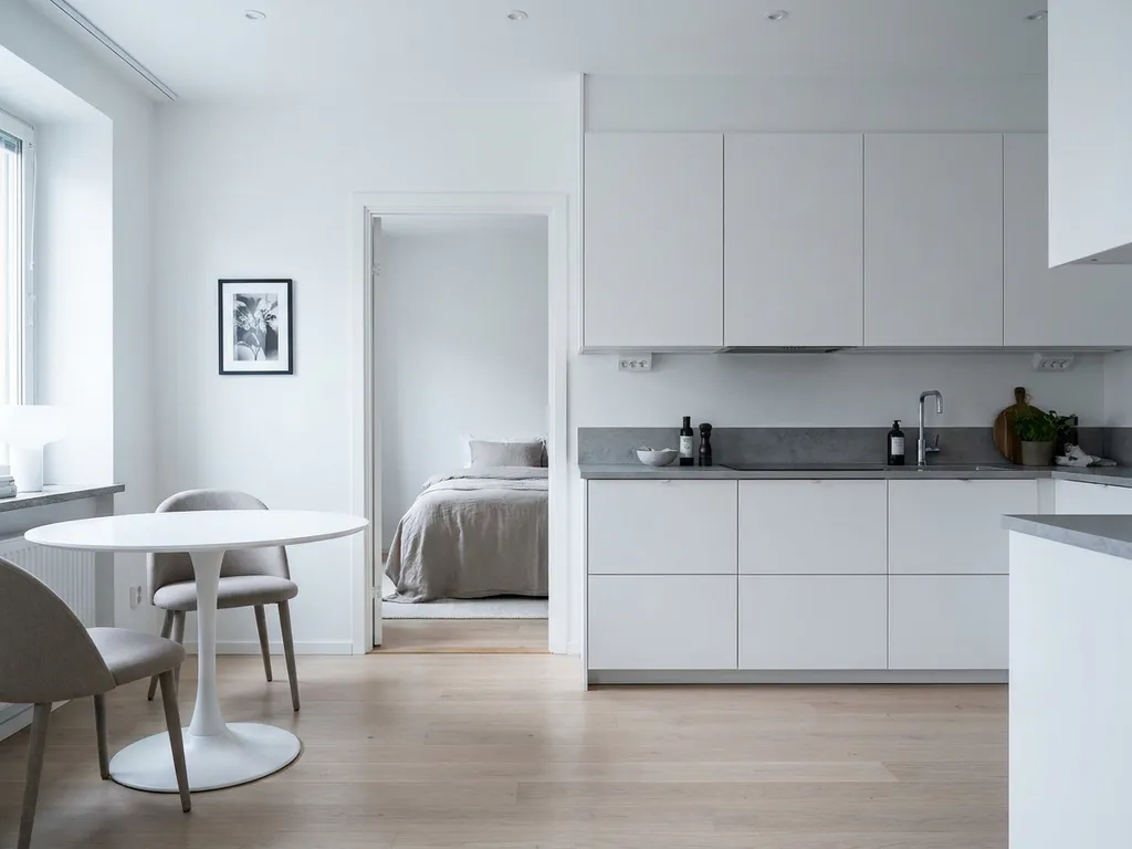 Minimalistisches Serviced Apartment mit hellem Holzboden, weißer Küche, grauer Arbeitsplatte und rundem Tulpen-Esstisch mit zwei greigen Stühlen.