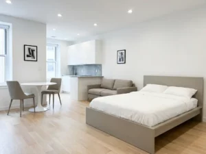 Minimalistisch eingerichtetes Apartment mit greigem Bettgestell, Zweisitzer-Sofa, rundem Tulpentisch und hellem Parkettboden.