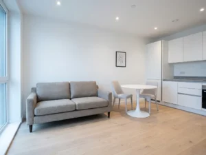Minimalistisch eingerichtete Wohnung mit hellem Parkettboden, weißer Einbauküche, Greigesofa und rundem Tulpen-Esstisch.