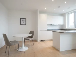 Minimalistisches Apartment mit hellem Parkettboden, weißer Einbauküche, grauer Steinarbeitsplatte und rundem Tulpen-Esstisch mit zwei greigen Polsterstühlen.