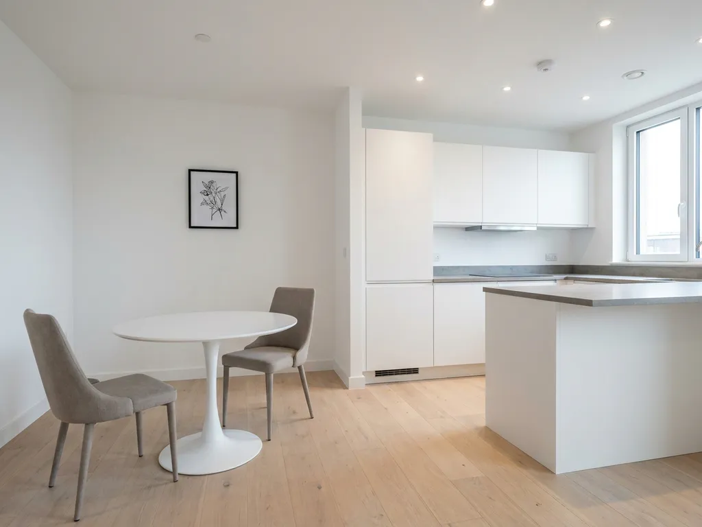 Minimalistisches Apartment mit hellem Parkettboden, weißer Einbauküche, grauer Steinarbeitsplatte und rundem Tulpen-Esstisch mit zwei greigen Polsterstühlen.