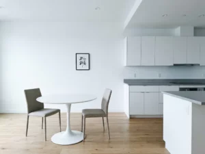 Minimalistisches Apartment mit hellem Parkettboden, weißer Einbauküche, grauer Steinarbeitsplatte und rundem Tulpen-Esstisch mit greigen Polsterstühlen.