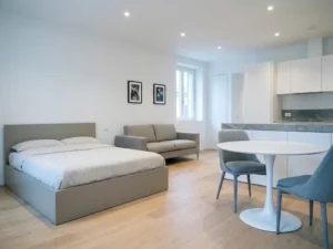 Minimalistisches Apartment mit greigefarbenem Bett, Zweisitzer-Sofa, rundem Tulpen-Esstisch und hellen Holzdielen.