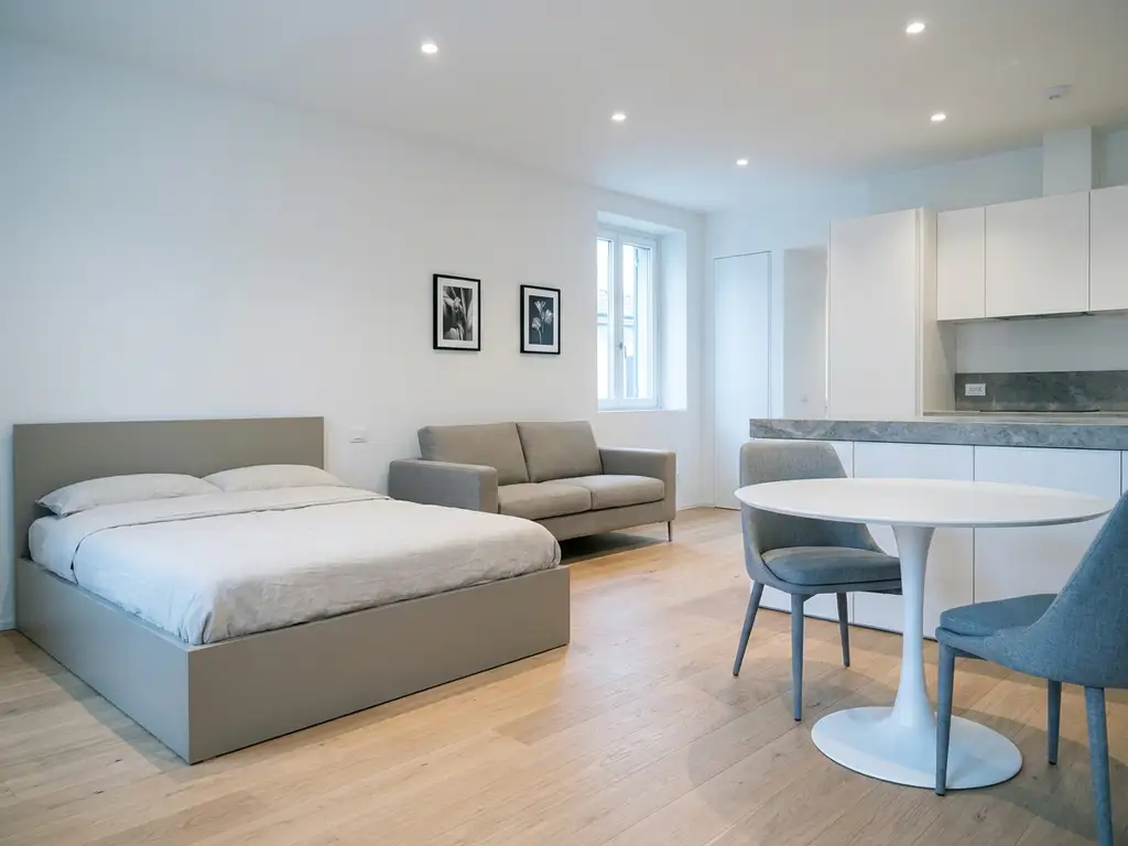Minimalistisches Apartment mit greigefarbenem Bett, Zweisitzer-Sofa, rundem Tulpen-Esstisch und hellen Holzdielen.