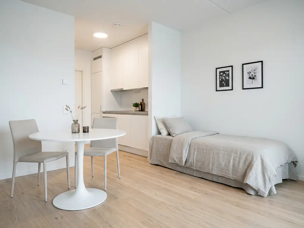 Minimalistisch eingerichtetes Apartment mit Einzelbett, rundem Tulpentisch und weißer Einbauküche auf hellem Eichenparkett.