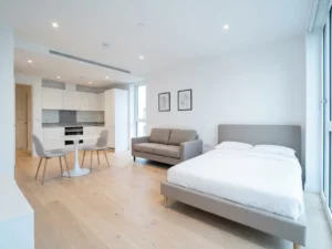 Minimalistisch eingerichtetes Apartment mit greigem Bettgestell, Zweisitzer-Sofa, rundem Tulpen-Esstisch und weißer Einbauküche auf hellem Parkettboden.