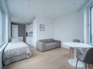 Minimalistisch eingerichtetes Apartment mit graubeigem Einzelbett, Sofa, rundem Tulpen-Esstisch und hellen Holzdielen.