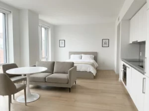 Minimalistisch eingerichtete Wohnung mit greigem Bett, Sofa, rundem Tulpentisch und hellen Holzböden in neutraler Farbpalette.