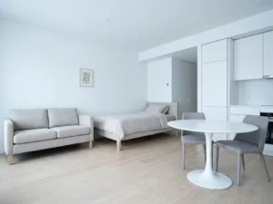 Minimalistisch eingerichtetes Apartment mit greigem Bett, weißem Tulpen-Esstisch und hellen Holzböden.