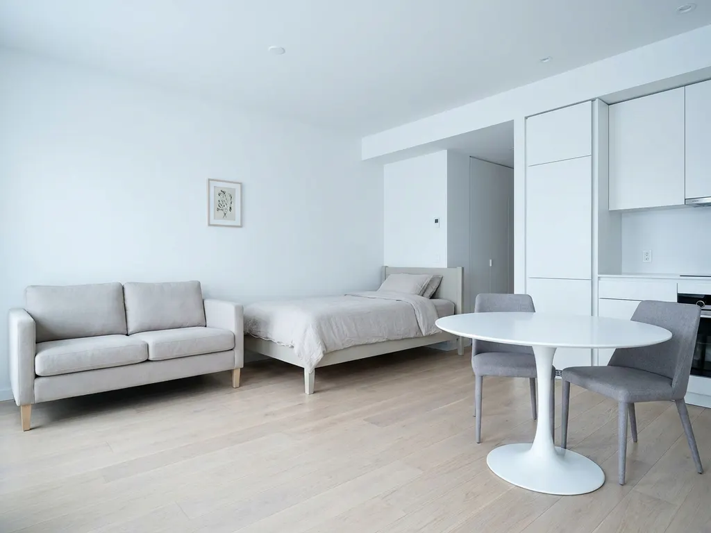 Minimalistisch eingerichtetes Apartment mit greigem Bett, weißem Tulpen-Esstisch und hellen Holzböden.